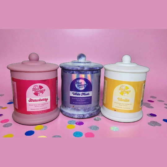Three scented candles with colorful labels on a pink background with confetti.