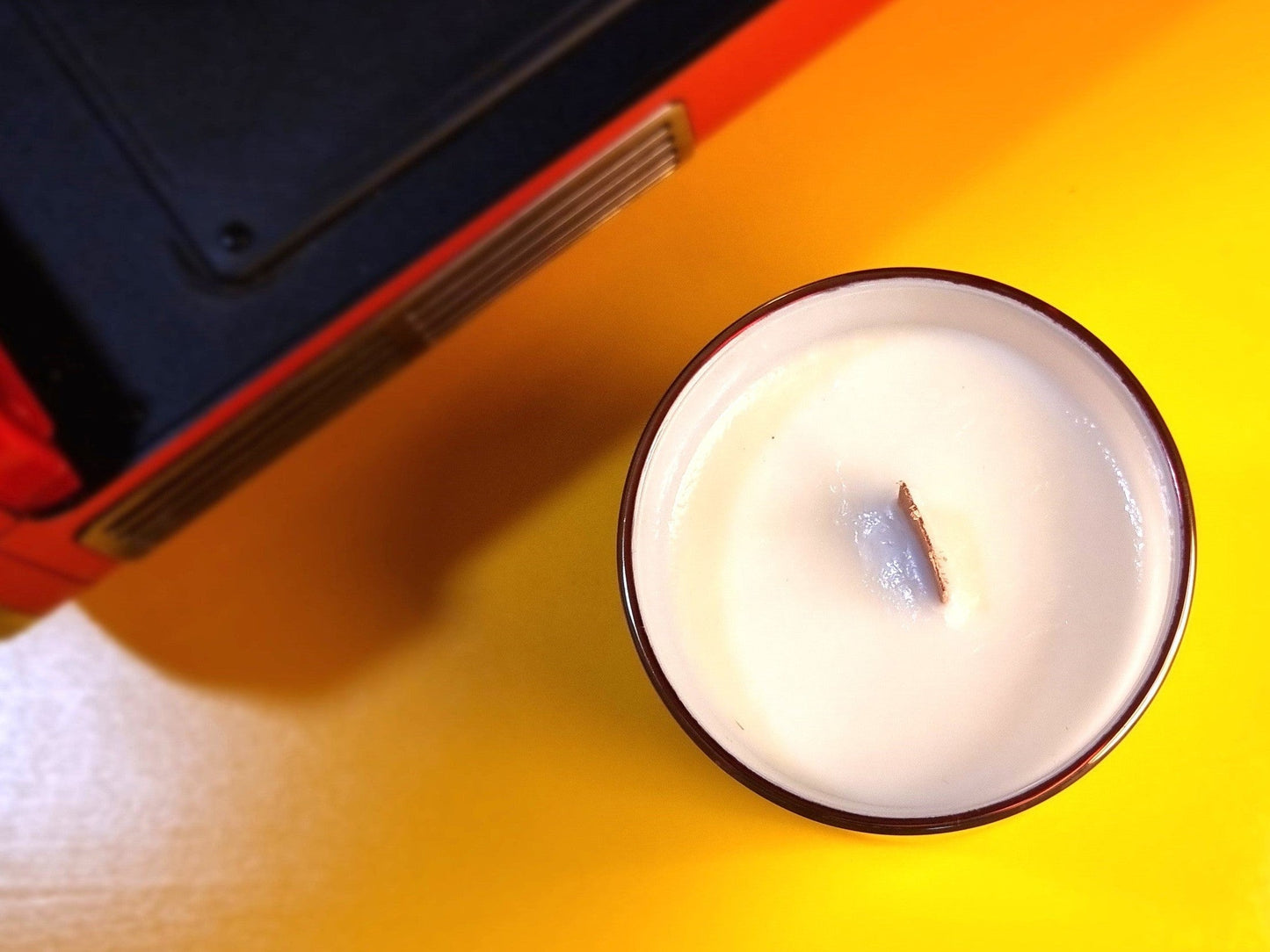 Candle in a white container on a yellow surface with a black object in the background