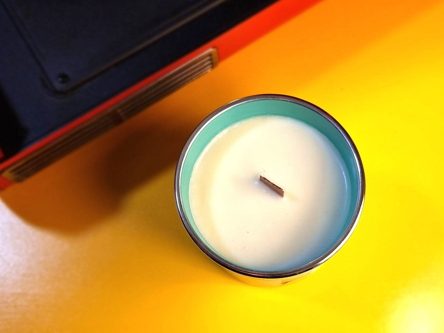 Candle in a glass holder on a yellow surface with a black object partially visible.