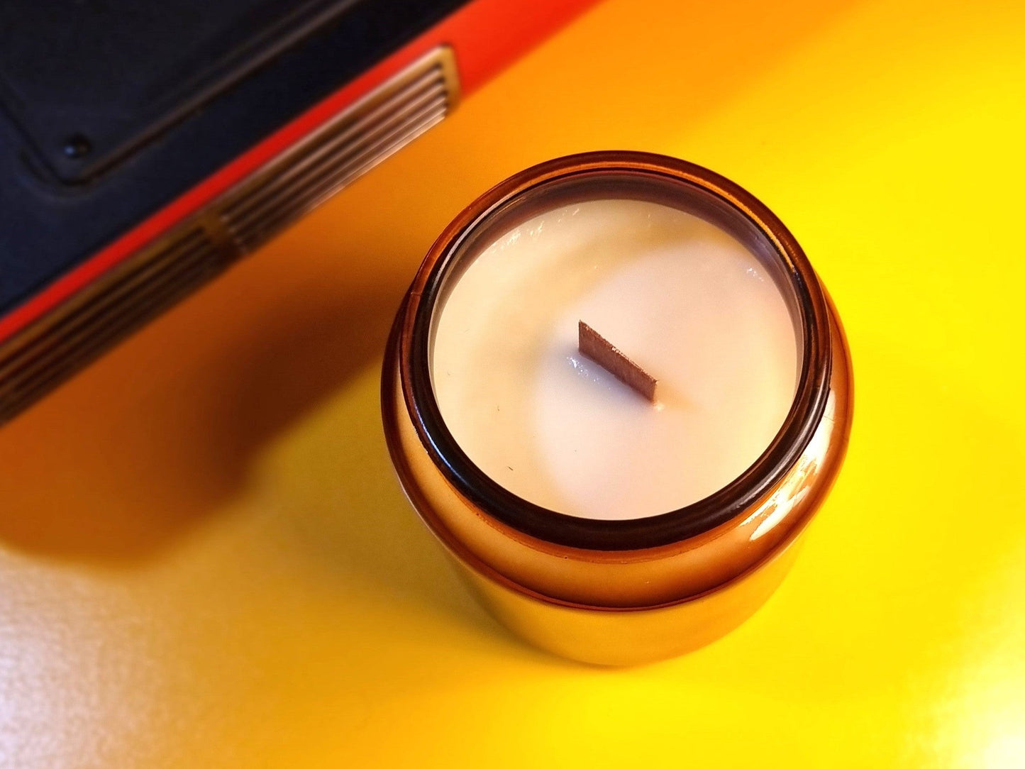 Candle in an amber jar on a yellow surface with a red and black object in the background