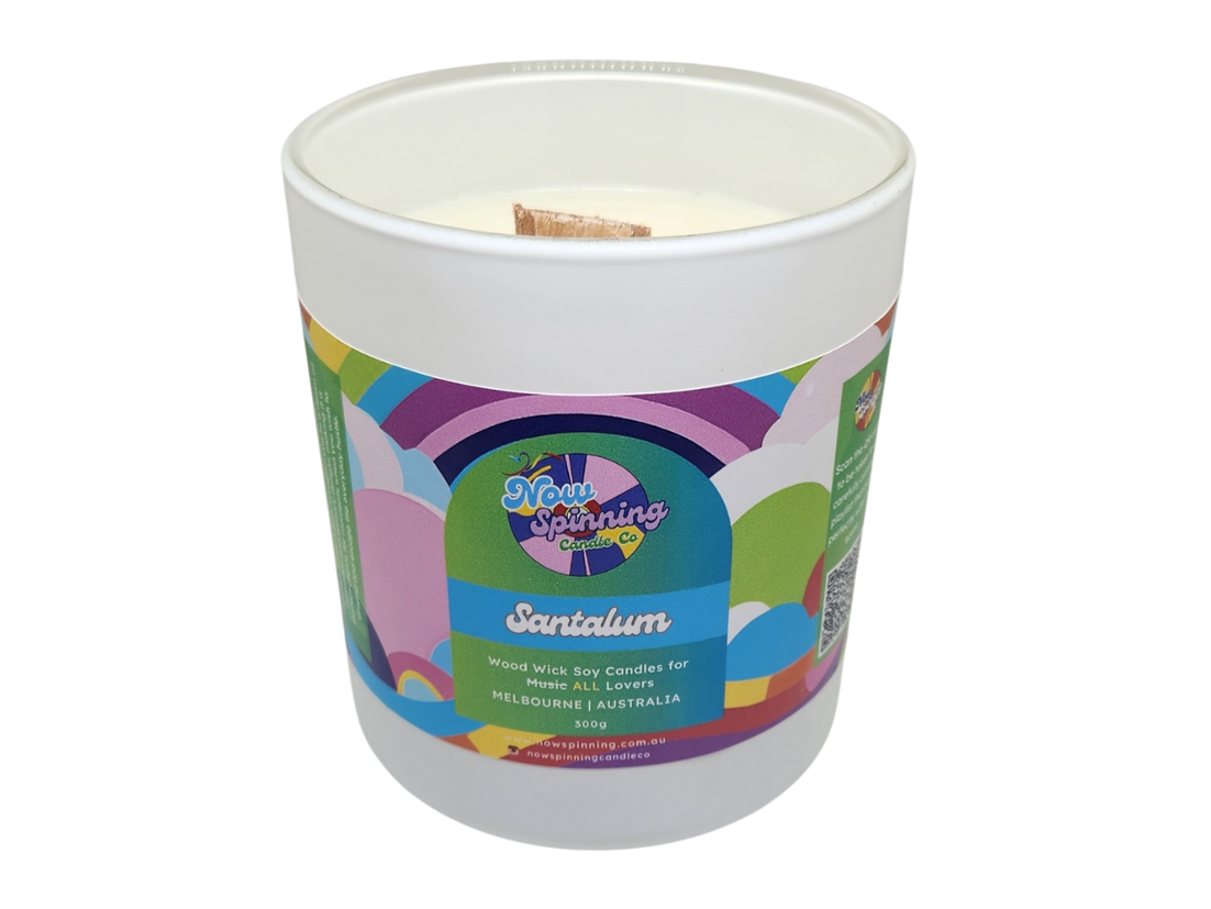 A white jar candle with a cork lid, labeled 'Santalum' by Now Spinning Candle Co with colorful patterns and text on the label.