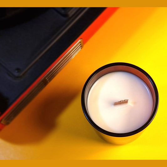 Candle in a metallic container on a yellow surface with a black and red object partially visible.