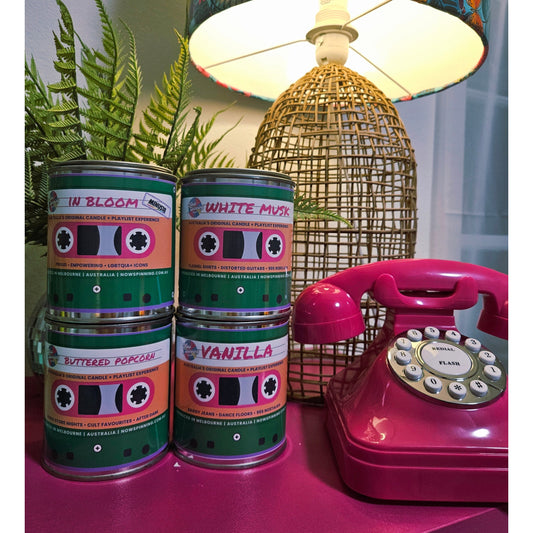 Four cans of Vanilla Bean Coffee Shop scented candles arranged on a pink surface next to a pink rotary telephone and a woven lamp base, with green foliage in the background.
