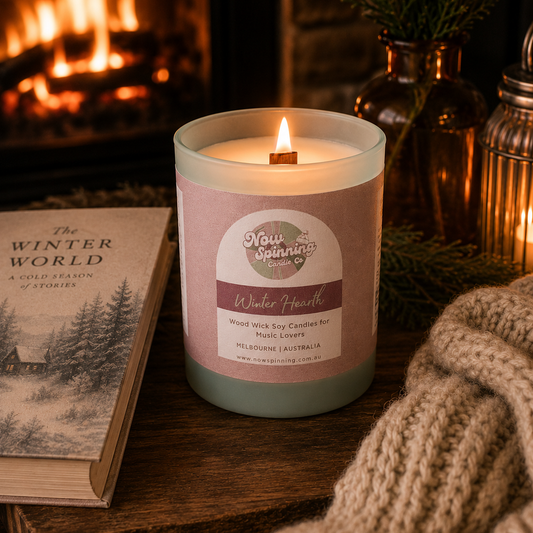 Winter Hearth wood wick soy candle by Now Spinning Candle Company displayed beside a book, chunky knit blanket and glowing fireplace. Featuring smoky firewood, amber and pine notes inspired by cosy winter evenings and fireside reading.