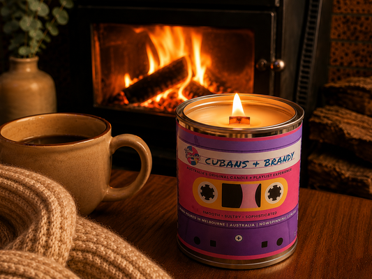 Now Spinning Cubans + Brandy candle with a glowing wood wick beside a coffee mug and fireplace.