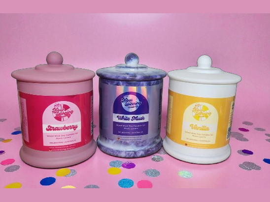 Three scented candles with colorful labels on a pink background with confetti.