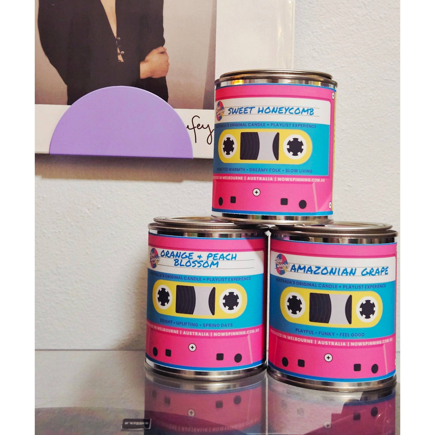 Three cans of Sweet Honeycomb perfume arranged in a pyramid on a reflective surface. The cans feature retro cassette tape graphics with pink, blue, yellow, and black colors, labeled with scents: Sweet Honeycomb, Orange + Peach Blossom, and Amazonian Grape.