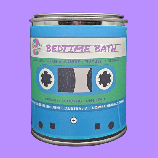 A cylindrical metal tin candle labeled 'BEDTIME BATH' with a cassette tape graphic on the front, shown against a solid lavender background.