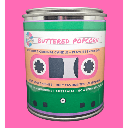 A novelty tin candle shaped like a cassette tape, labeled 'Buttered Popcorn' by Nowspinning, set against a solid pink background.