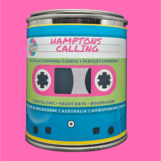 A novelty candle tin styled like a retro cassette tape, featuring a blue, green, pink, and yellow label with the text 'HAMPTONS CALLING' on a solid pink background.