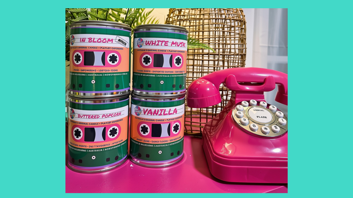 Four cans of coffee flavor pairs arranged in two stacks next to a retro pink rotary telephone on a pink surface, with a wicker basket and greenery in the background. The cans feature retro cassette tape labels with flavors including 'In Bloom', 'White Murk', 'Butterfly Papercake', and 'Vanilla'.