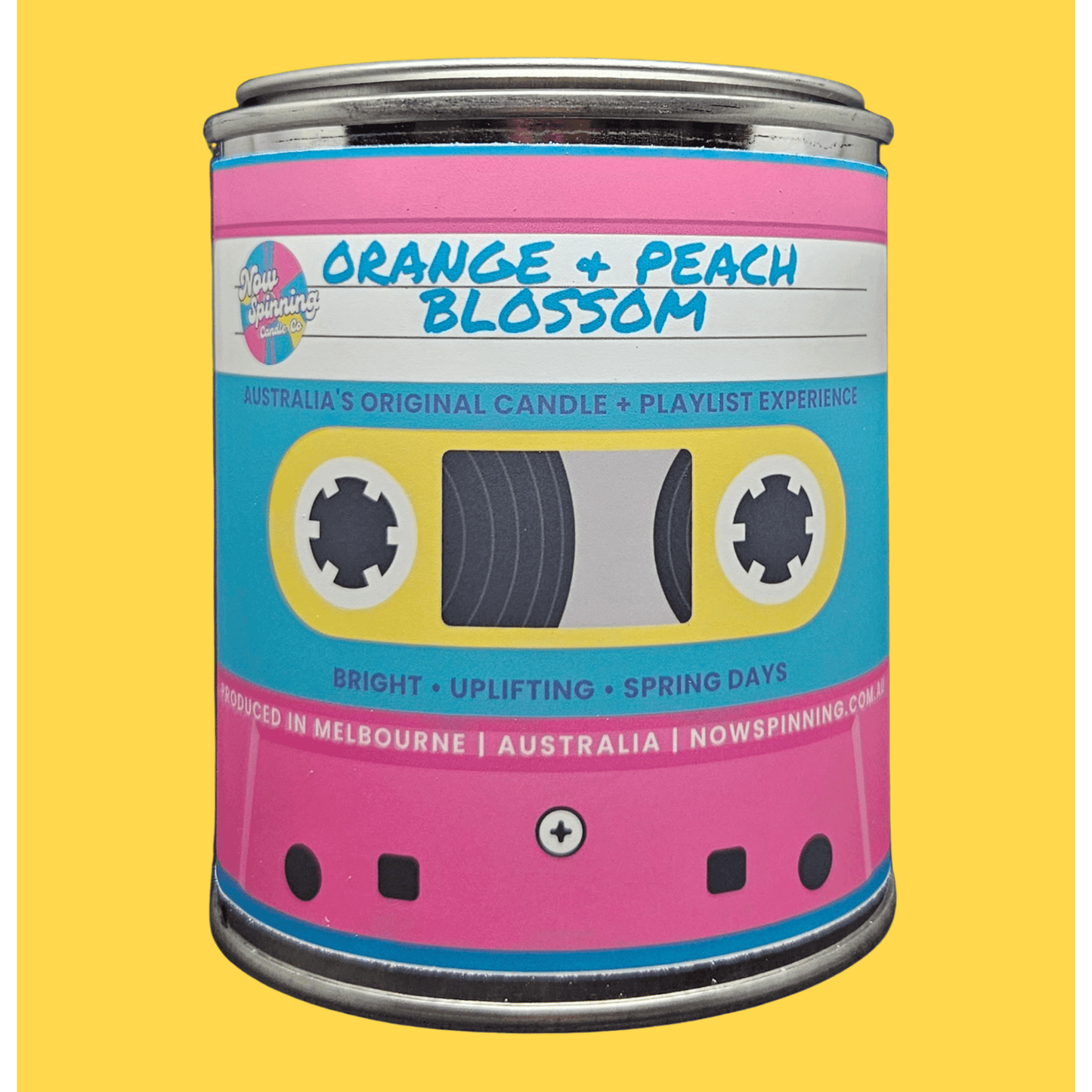 A novelty candle tin styled like a retro cassette tape, labeled 'Orange + Peach Blossom' by Nowspinning Co, set against a solid yellow background.