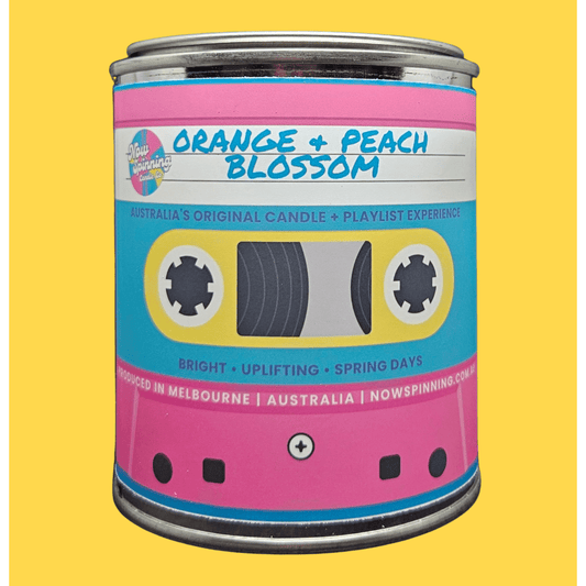 A novelty candle tin styled like a retro cassette tape, labeled 'Orange + Peach Blossom' by Nowspinning Co, set against a solid yellow background.