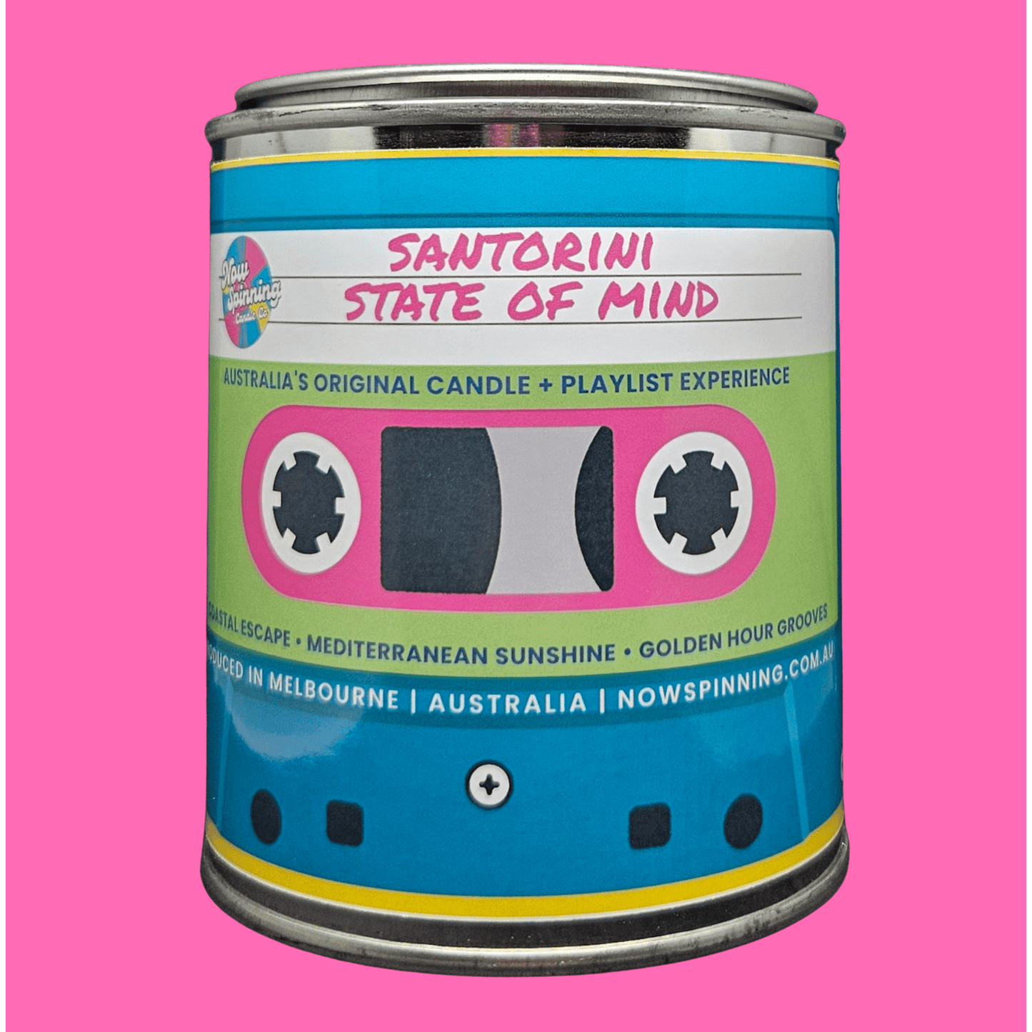 A cylindrical metal tin candle labeled 'SANTORINI STATE OF MIND' with a bright blue, pink, and light green retro cassette-tape style label, shown on a solid pink background.