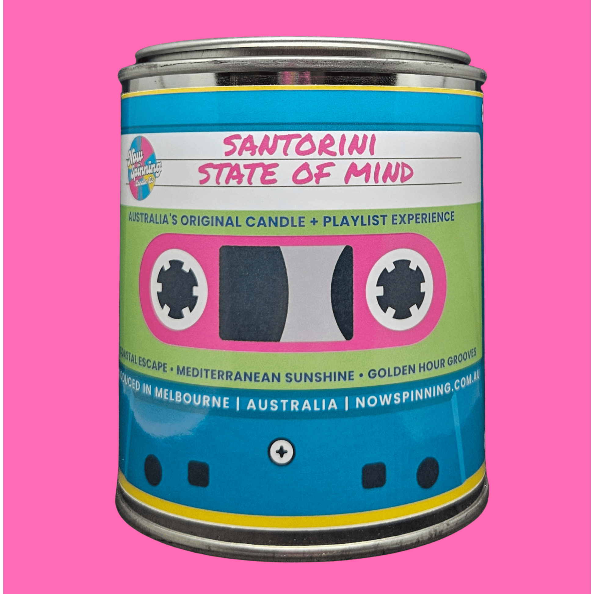 A cylindrical metal tin candle labeled 'SANTORINI STATE OF MIND' with a bright blue, pink, and light green retro cassette-tape style label, shown on a solid pink background.