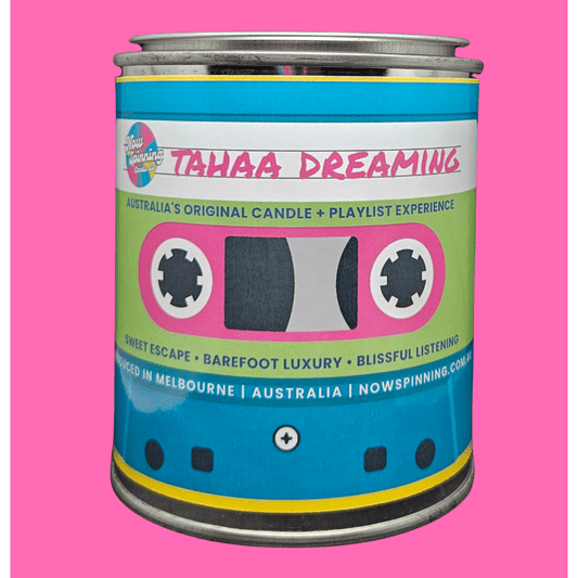 A novelty candle tin styled like a cassette tape, featuring a bright blue and lime green label with pink and white details, set against a solid pink background.