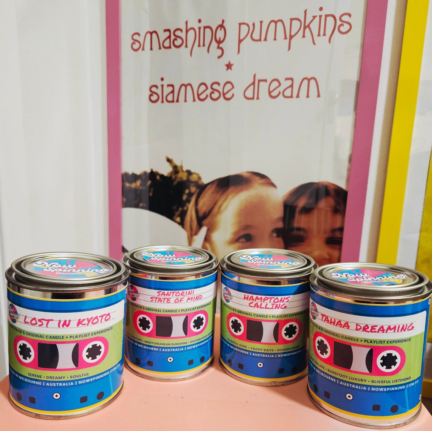 Four metal paint cans with colorful cassette-tape-style labels are lined up on a pink surface in front of a framed print that reads 'smashing pumpkins siamese dream'.
