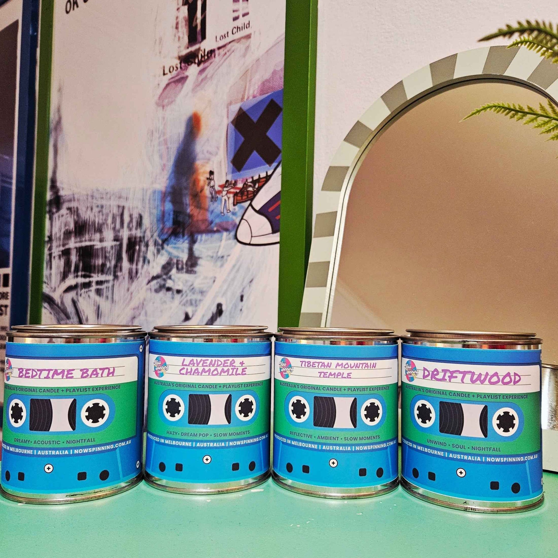 Four decorative tin cans with cassette-tape-style labels are lined up on a mint green surface. The labels read 'BEDTIME BATH', 'LAVENDER + CHAMOMILE', 'TIBETAN MOUTAIN TEMPLE', and 'DRIFTWOOD'.