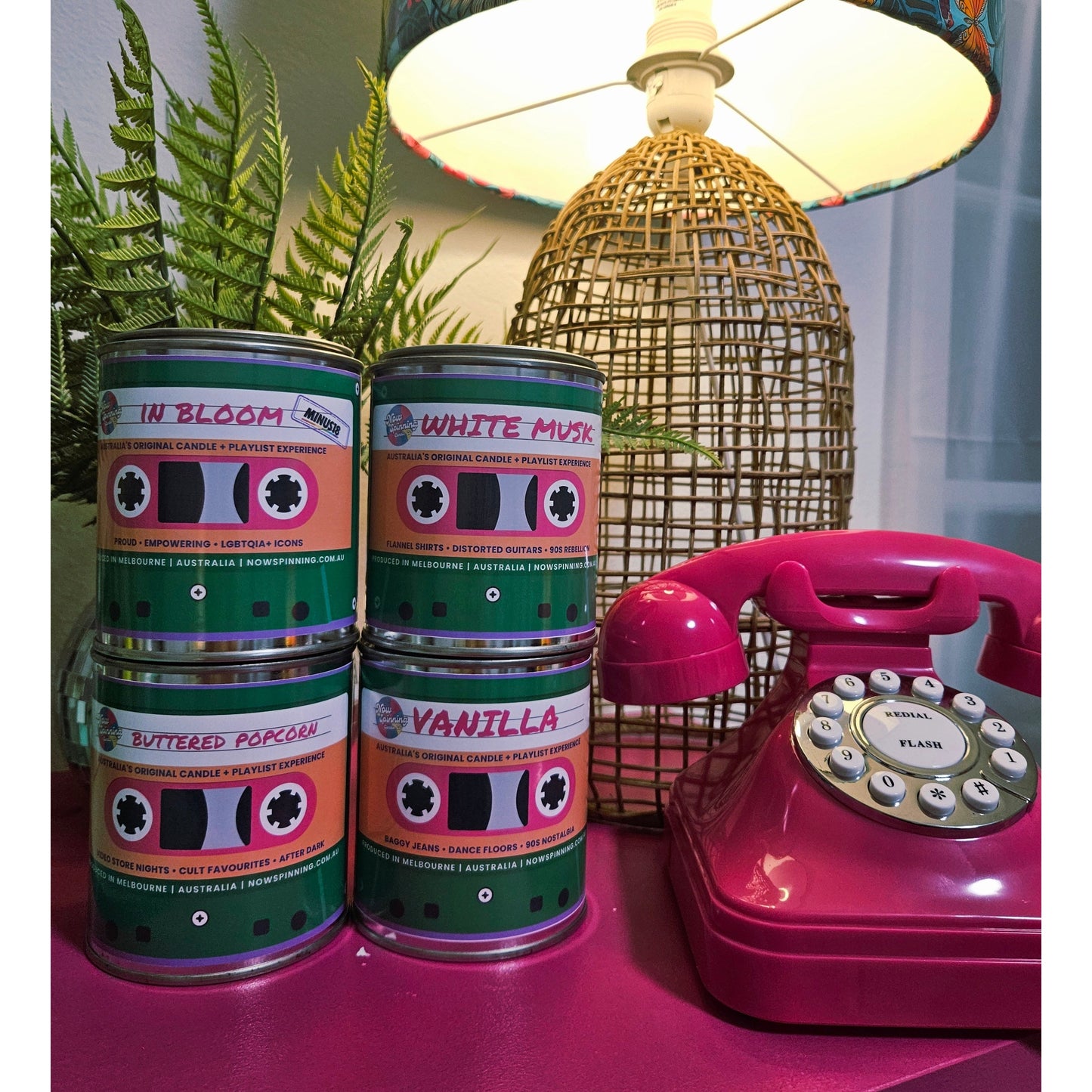 Four cans of Vanilla Bean Coffee Shop scented candles arranged on a pink surface next to a pink rotary telephone and a woven lamp base, with green foliage in the background.