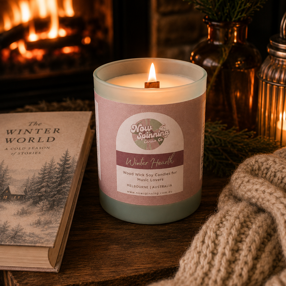 Winter Hearth wood wick soy candle by Now Spinning Candle Company displayed beside a book, chunky knit blanket and glowing fireplace. Featuring smoky firewood, amber and pine notes inspired by cosy winter evenings and fireside reading.